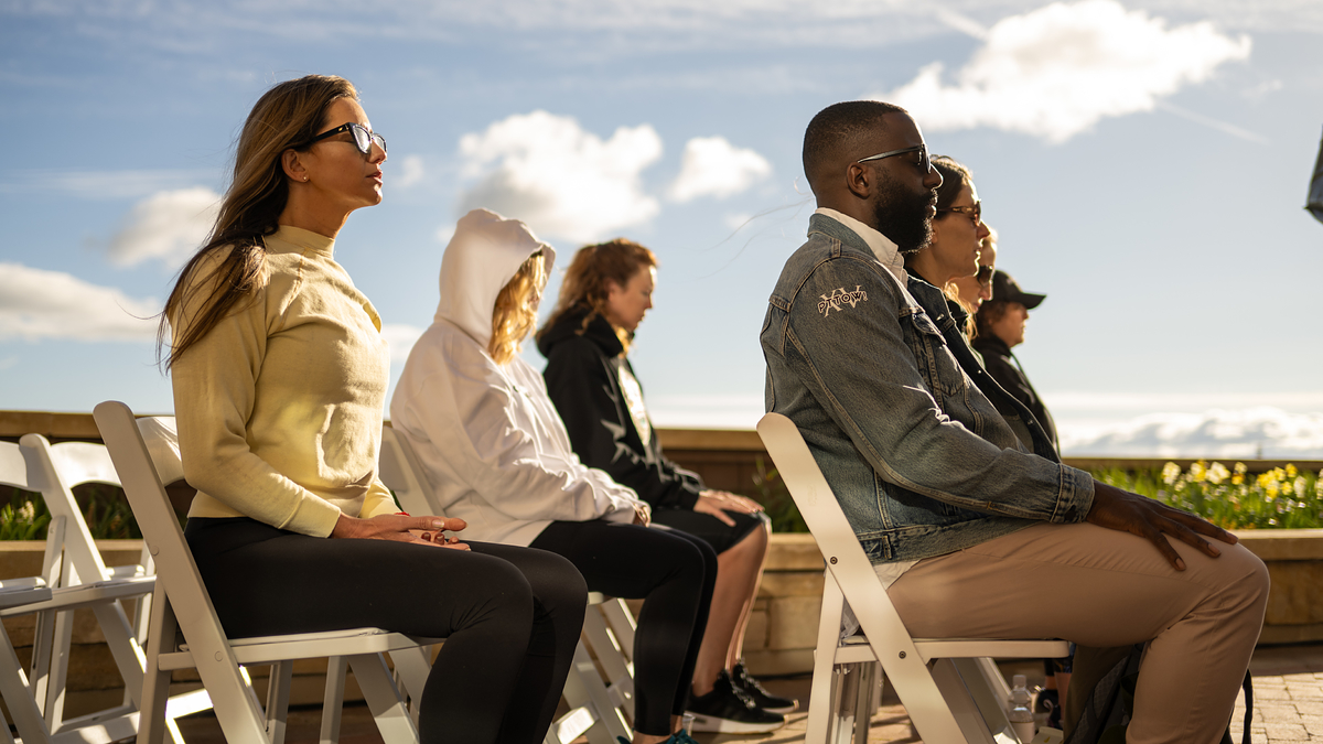 People sit on white chairs outdoors facing the same direction, wearing casual clothes and sunglasses in a serene setting with blue sky and clouds