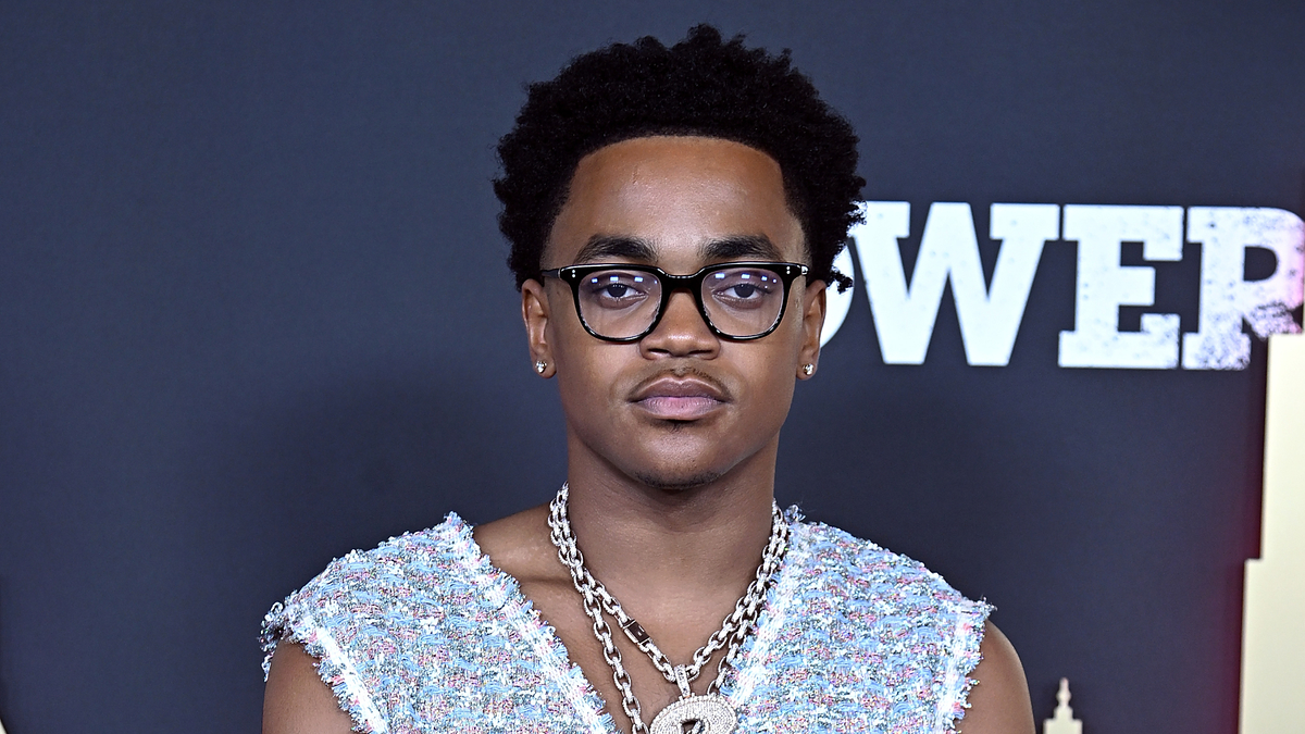 Michael Rainey Jr. on the red carpet, wearing a sleeveless, textured top and layered chain necklaces. Background features "Power" branding