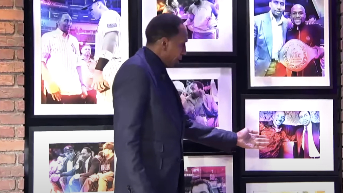 Stephen A. Smith pointing at a wall of framed photos featuring various moments with individuals, including one with a boxing champion belt