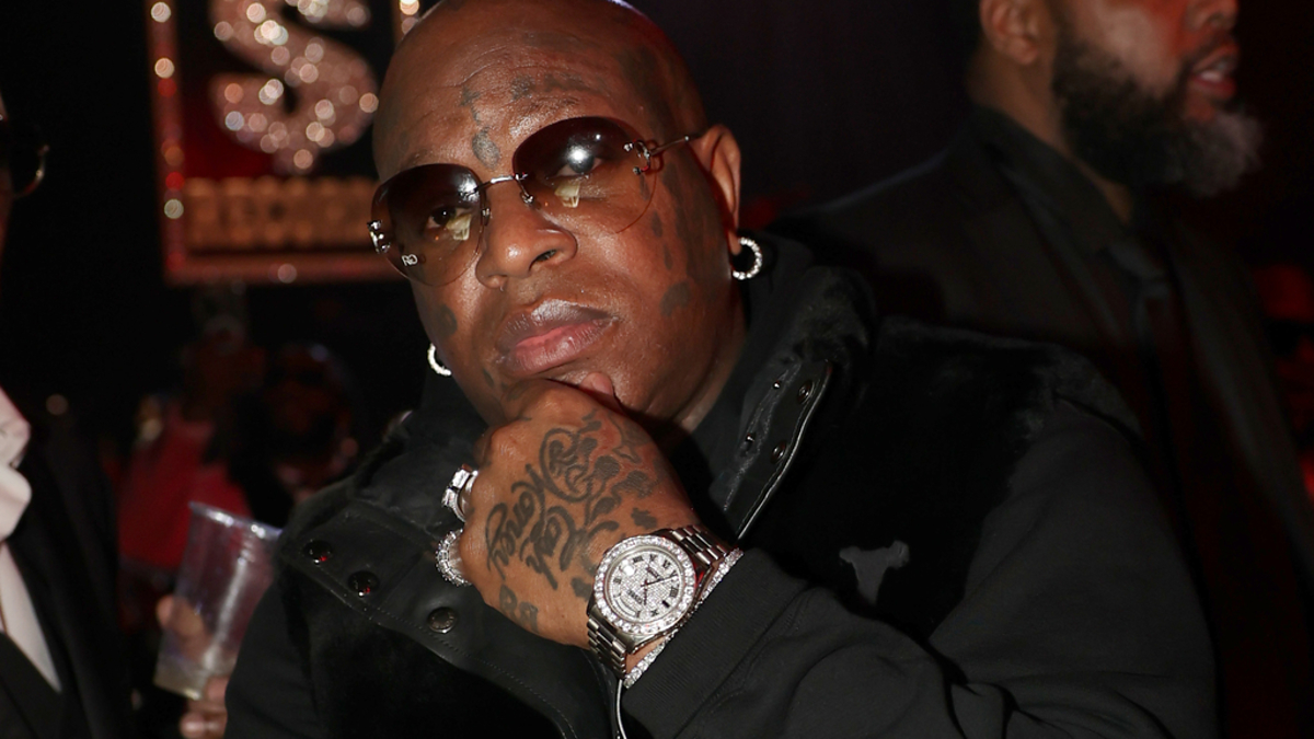 Birdman, wearing a black jacket and sunglasses, poses with his arms crossed while showing off his tattoos and jewelry at a music event