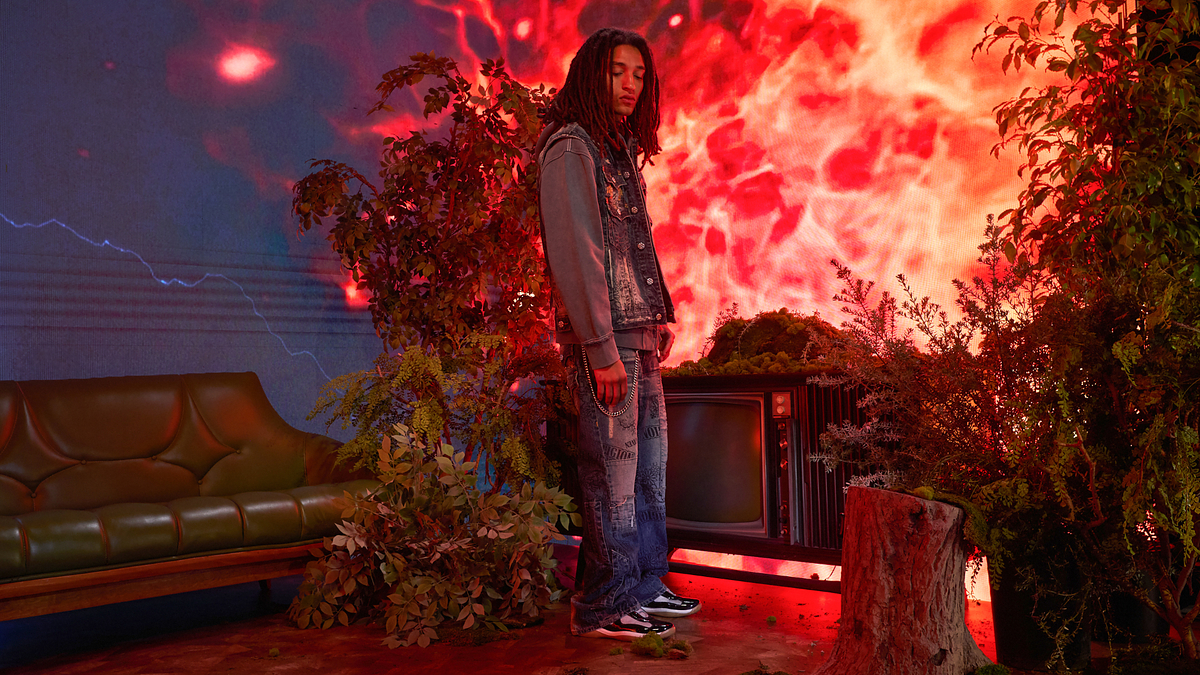 Person wearing denim jacket and jeans stands next to foliage, vintage TV, and log in a staged set with intense red backdrop