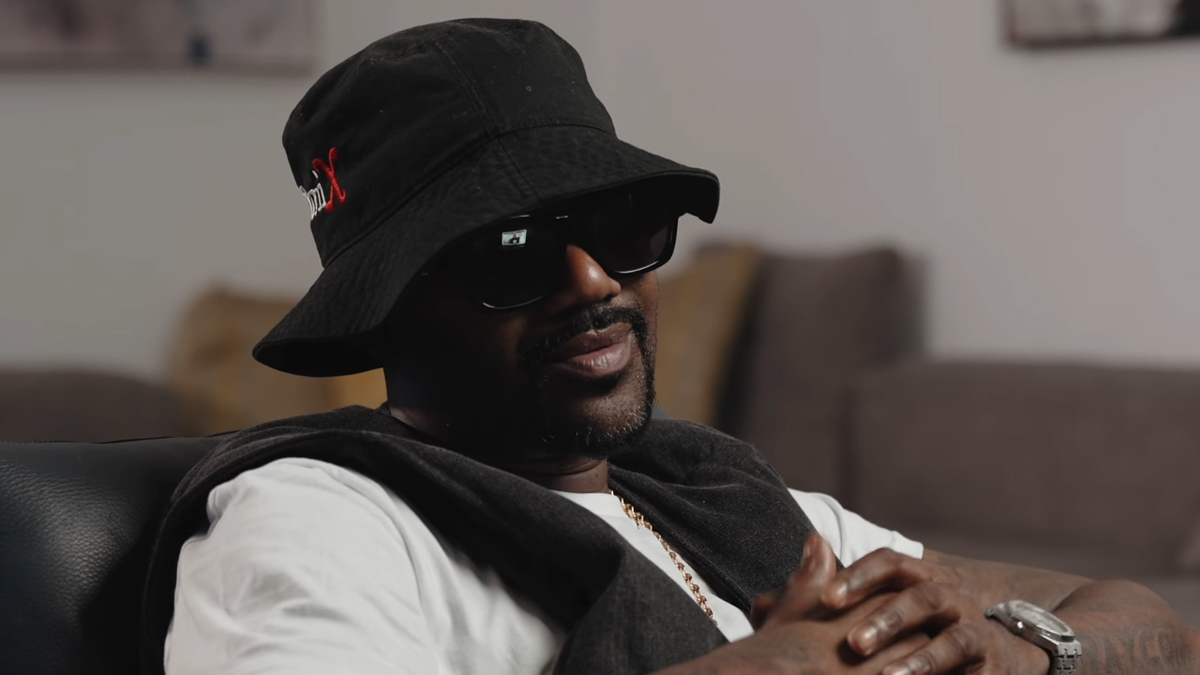 A man sits wearing a black bucket hat, dark sunglasses, and a white shirt with a dark jacket draped over his shoulders