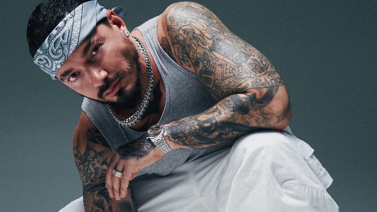 J Balvin crouches down, wearing a bandana, a sleeveless top, and white pants. His arms are covered in tattoos and he is accessorized with multiple necklaces and a watch