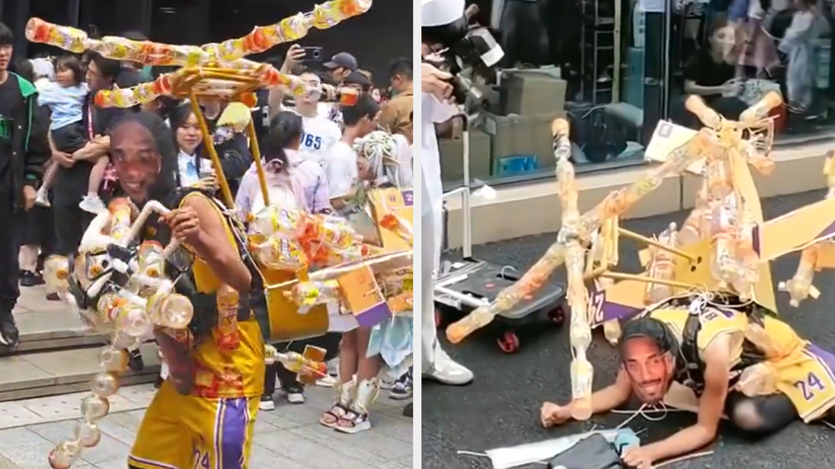 An individual cosplaying as Kobe Bryant wearing the late player's Lakers face on a ski mask and a makeshift helicopter suit made out of cardboard and plastic bottles. He lays on the sidewalk as a forensic photographer takes photos of him.