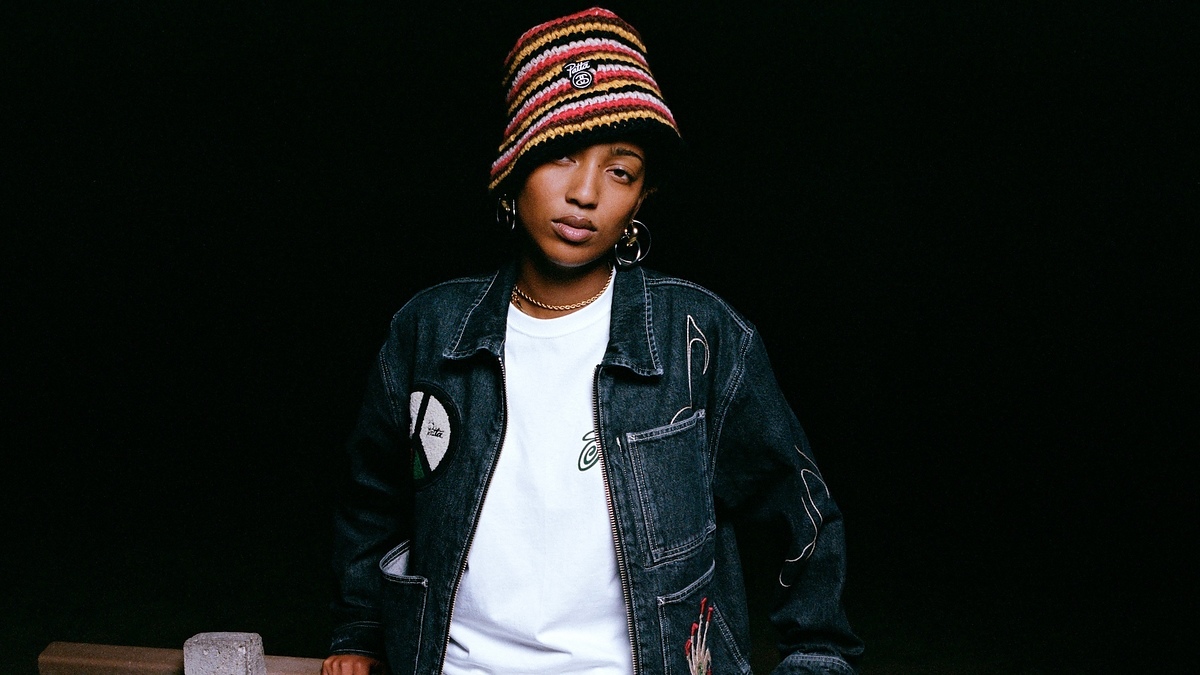 A person wearing a multicolored knit hat, a denim jacket with patches, hoop earrings, and a white shirt stands against a dark background