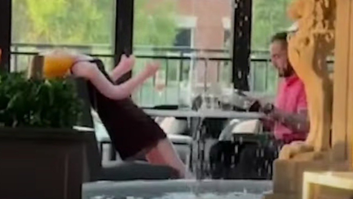 A person reads a menu while a mannequin dressed in dark clothing dramatically falls backward into a water feature at an outdoor dining area