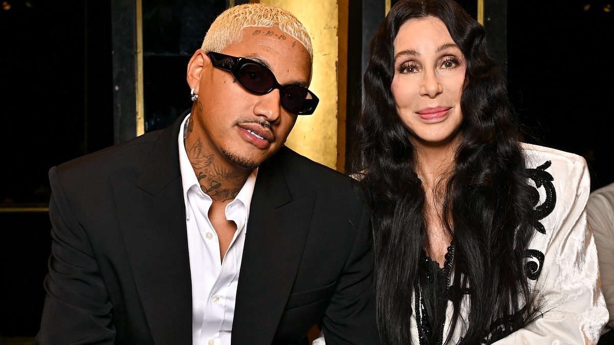 Cher and Alexander Edwards are seated together, with Cher wearing a stylish white jacket adorned with black details and Edwards in a black suit and sunglasses