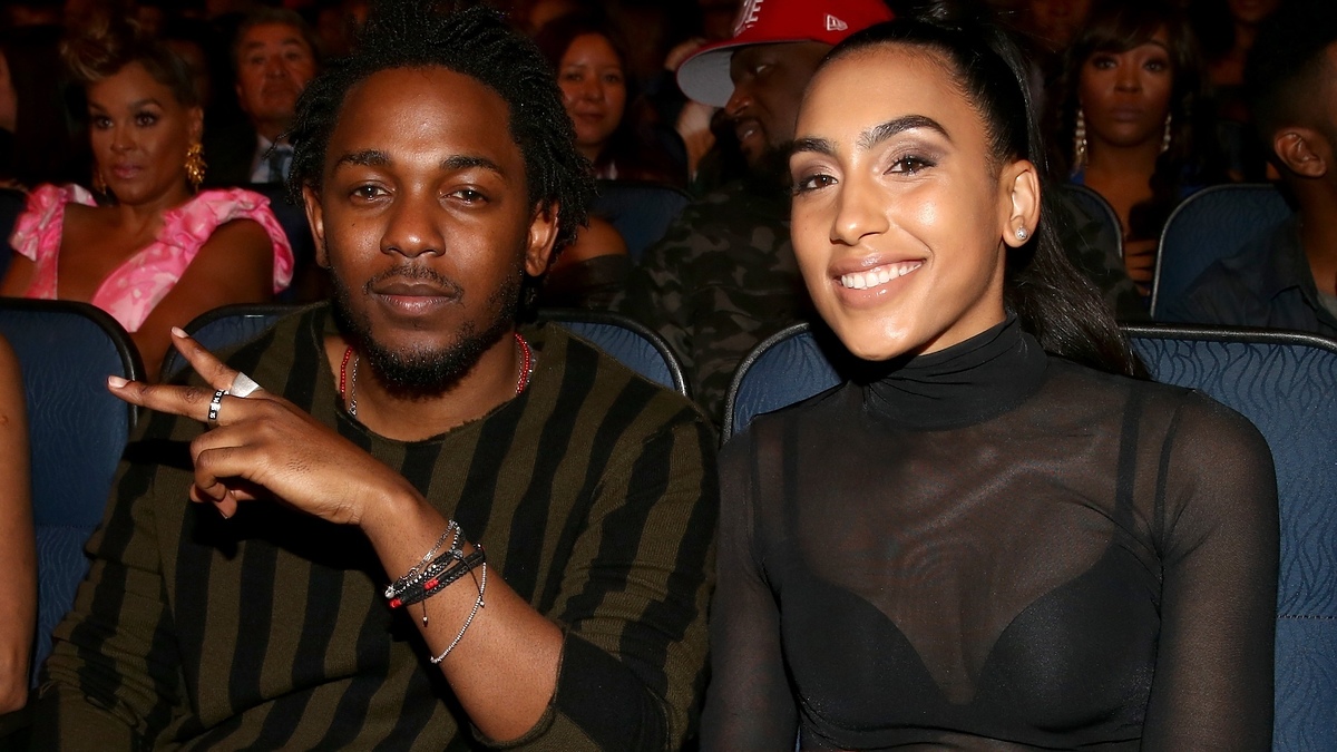 Kendrick Lamar, in a striped sweater, and Whitney Alford, in a sheer black top, sit closely together at an event, smiling at the camera