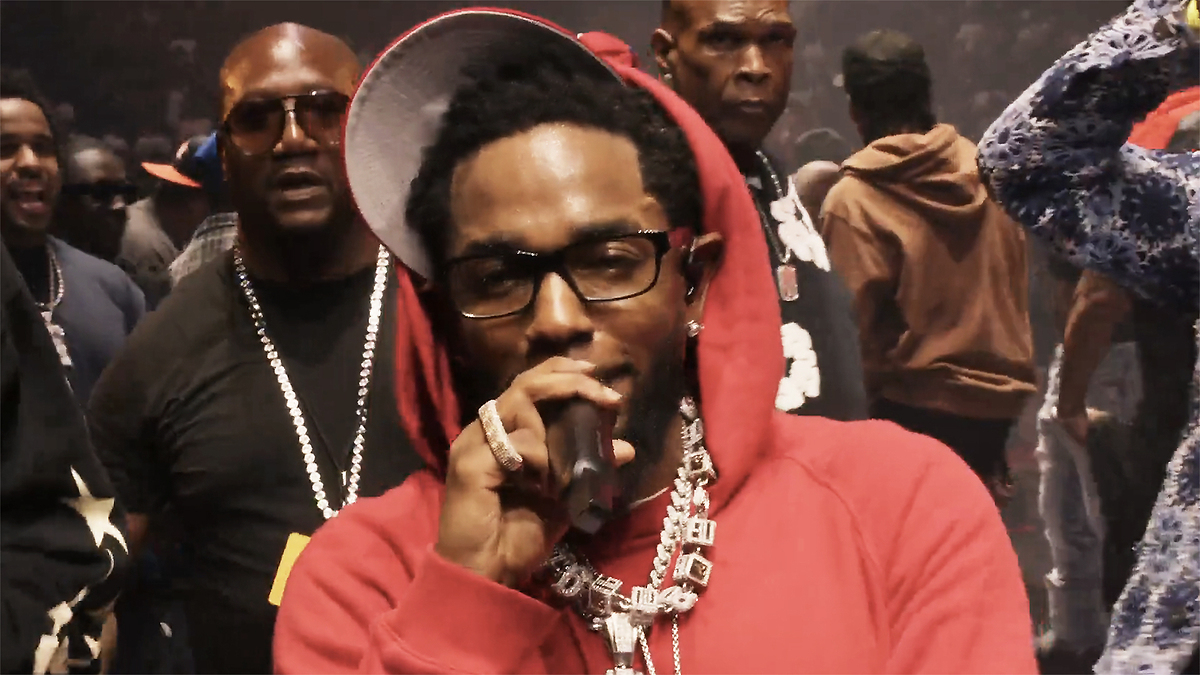 Man wearing glasses and a hoodie sings into a microphone, surrounded by others in a crowded setting