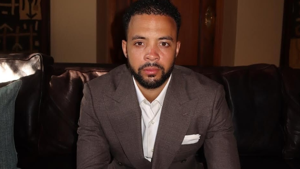 A man with a beard, wearing a brown suit and white shirt, sits on a couch, looking directly at the camera