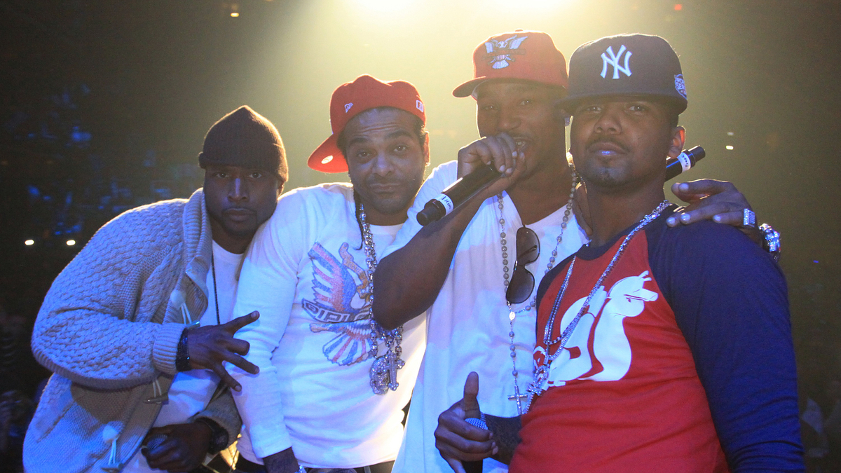 Cam'ron, Jim Jones, Juelz Santana, and Freekey Zekey on stage, wearing casual and urban-style clothes, with baseball caps and jewelry, posing for a photo