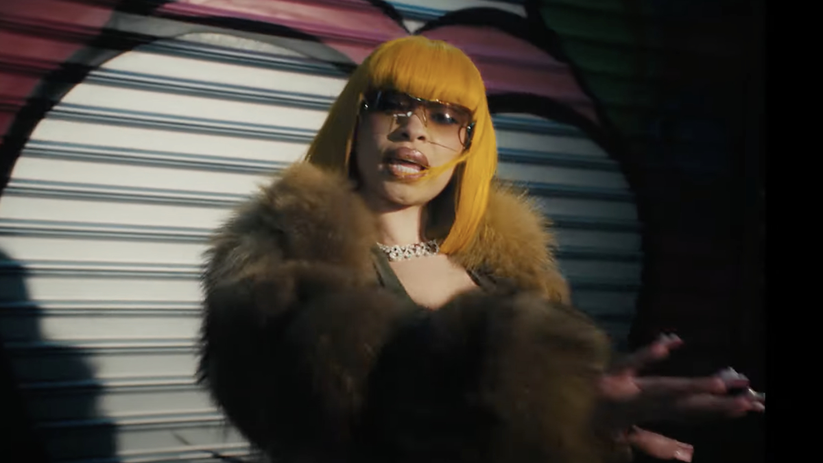 Ice Spice wears a fur coat and sunglasses, posing with her hands outstretched in front of what appears to be a metal shutter in a music video scene
