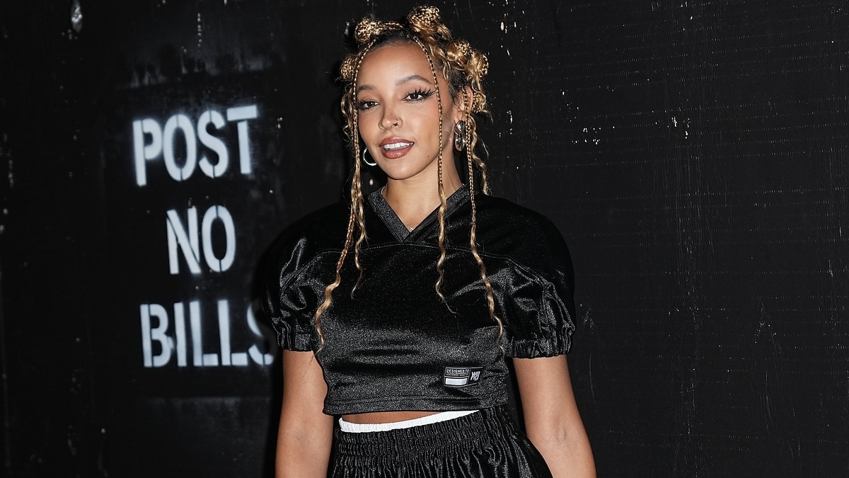 Tinashe standing in front of a wall with a "Post No Bills" sign, wearing a sporty black outfit with braided hair styled in Bantu knots