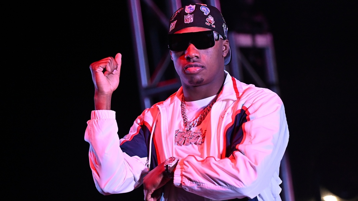 A man in a white tracksuit with stripes and a hat decorated with various pins, wearing sunglasses, strikes a pose with one arm raised.

