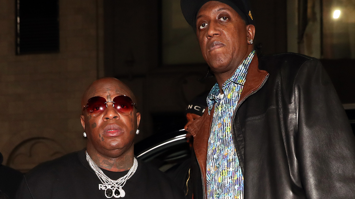 Birdman in a necklace with multiple diamond chains stands next to Slim dressed in a leather jacket and patterned shirt at a music event