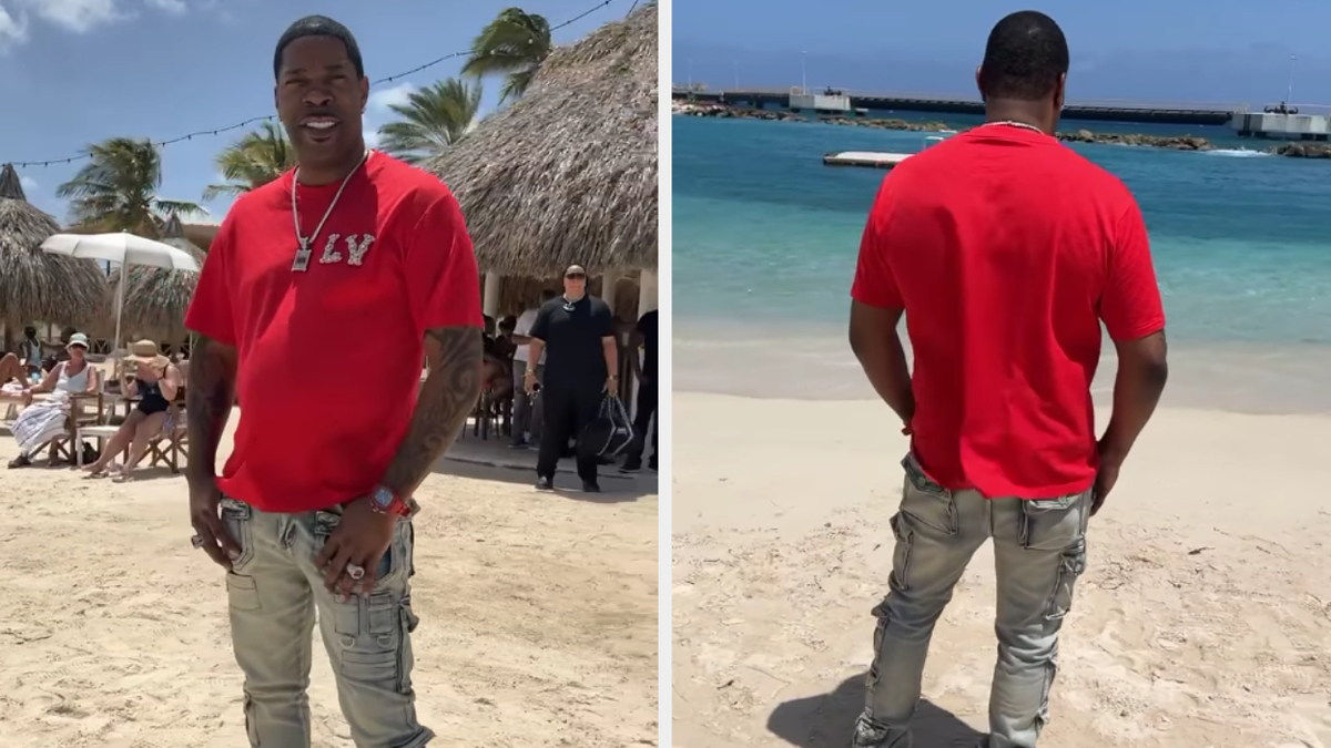 Busta Rhymes is on a beach in casual attire, wearing a red T-shirt and light ripped jeans. The ocean is visible in the background