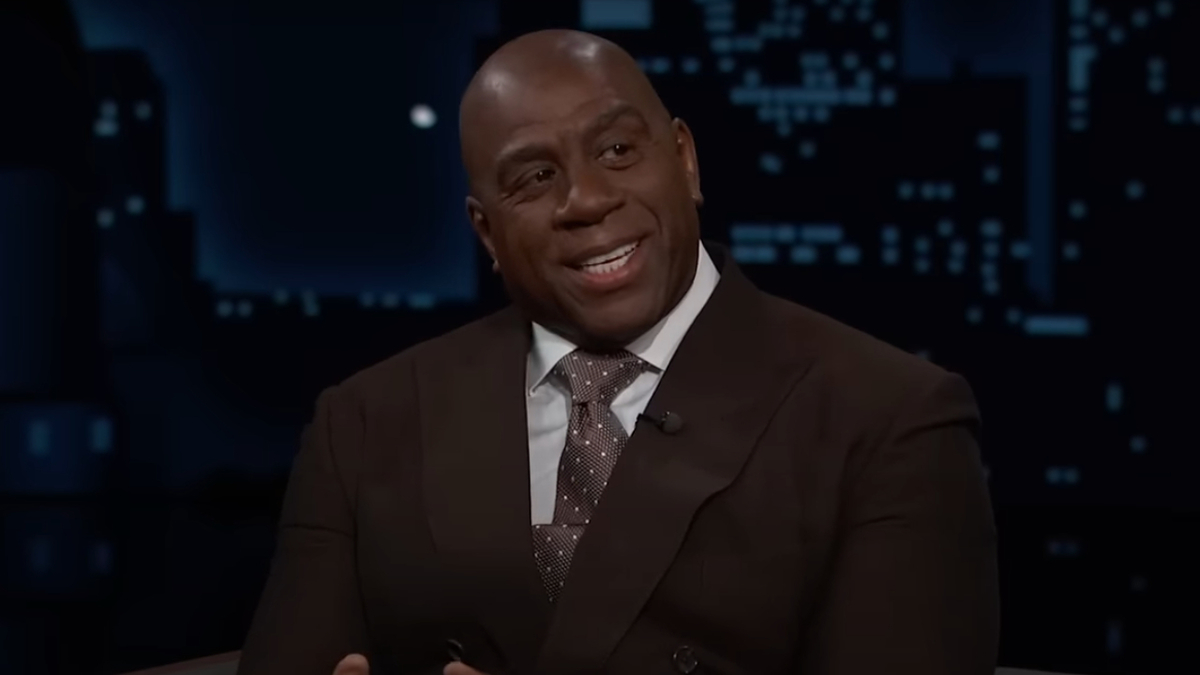 Magic Johnson in a brown suit and polka dot tie, smiling during a television interview