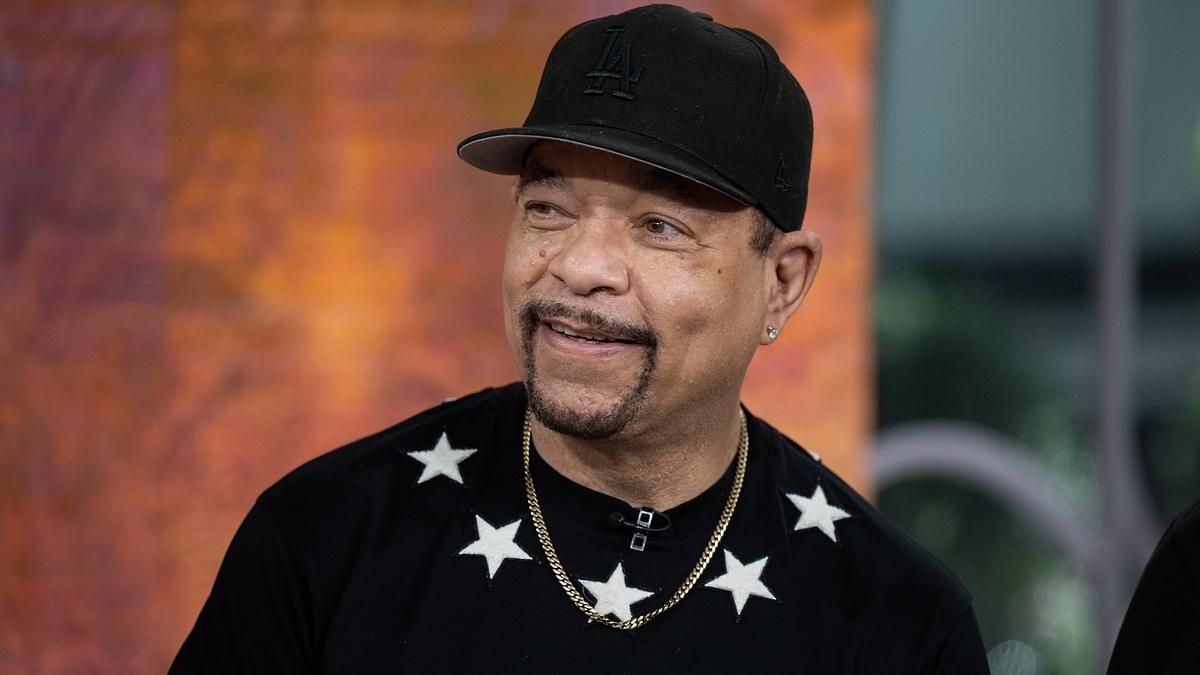 Ice-T wears a black cap and a black shirt with white stars, smiling while appearing on a show