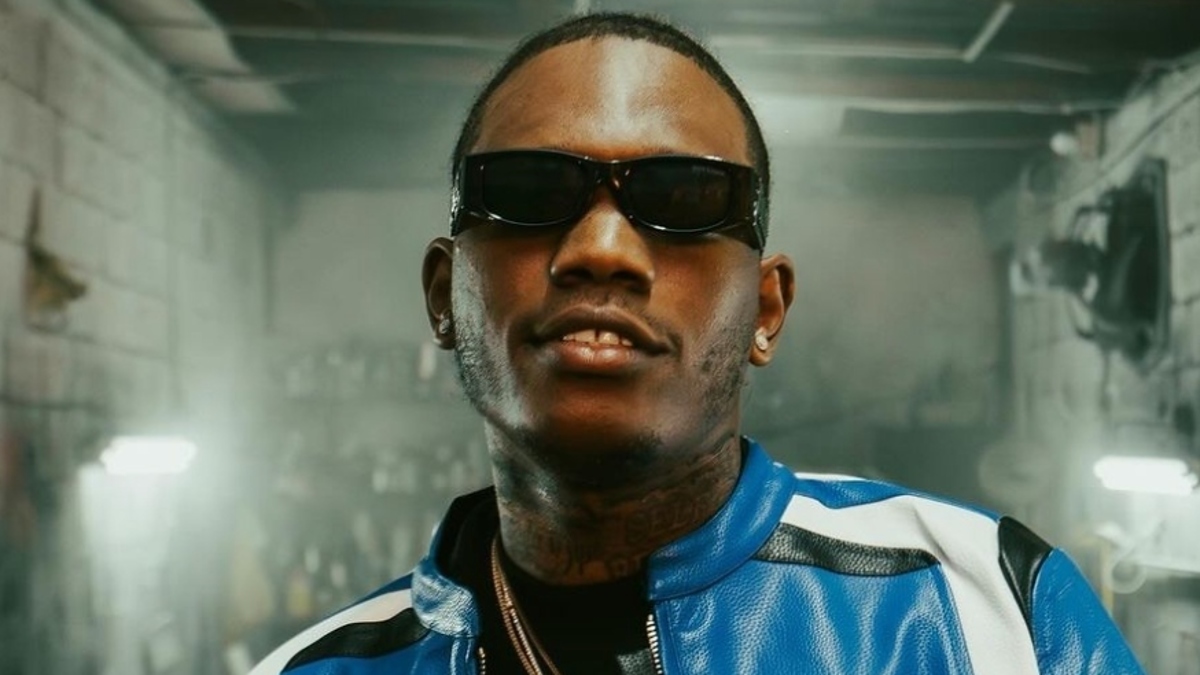 Rapper Hotboy Wes wearing sunglasses, a blue and white jacket, and layered necklaces, standing in an industrial-looking space