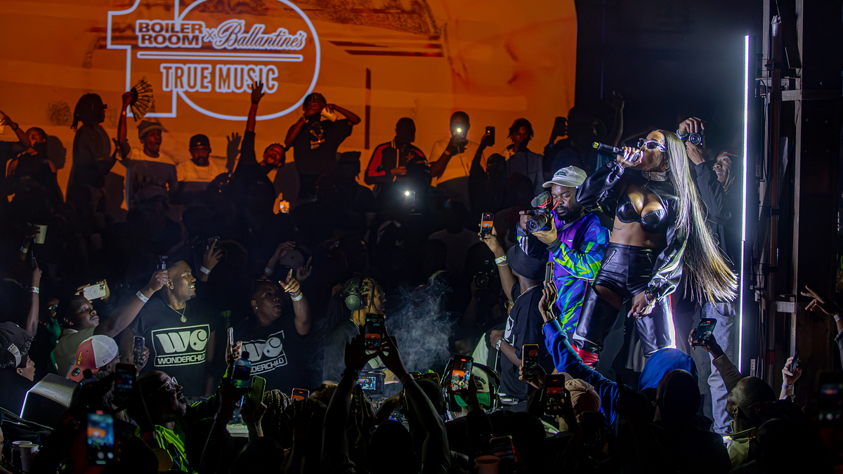 A performer in futuristic clothing sings on stage at a Boiler Room and Ballantine's True Music event, surrounded by a lively crowd taking pictures
