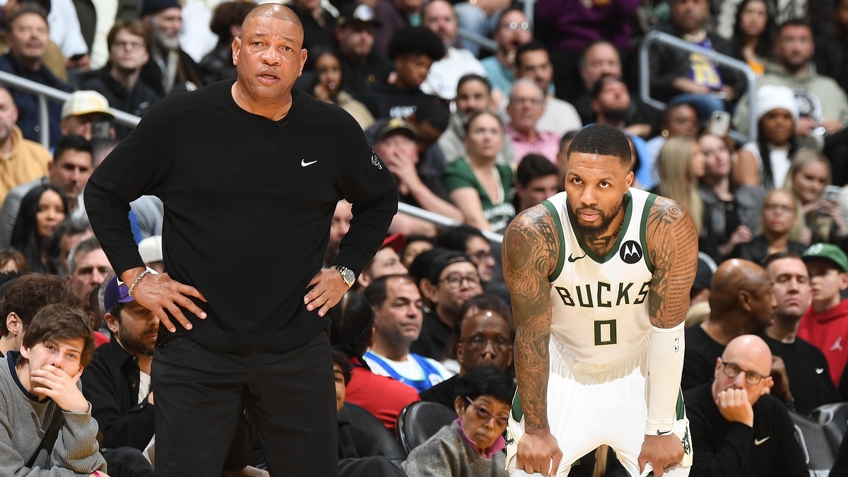 Doc Rivers (left) and Damian Lillard, wearing Bucks jersey #0, stand on the sidelines during a basketball game with a crowd watching in the background