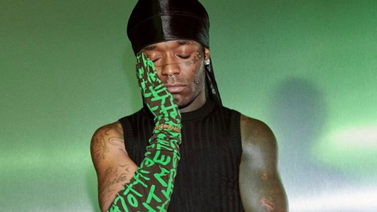 Lil Uzi Vert modeling for Marc Jacobs, wearing a black sleeveless top, black skirt, and boots, and holding a green graffiti-print tote bag