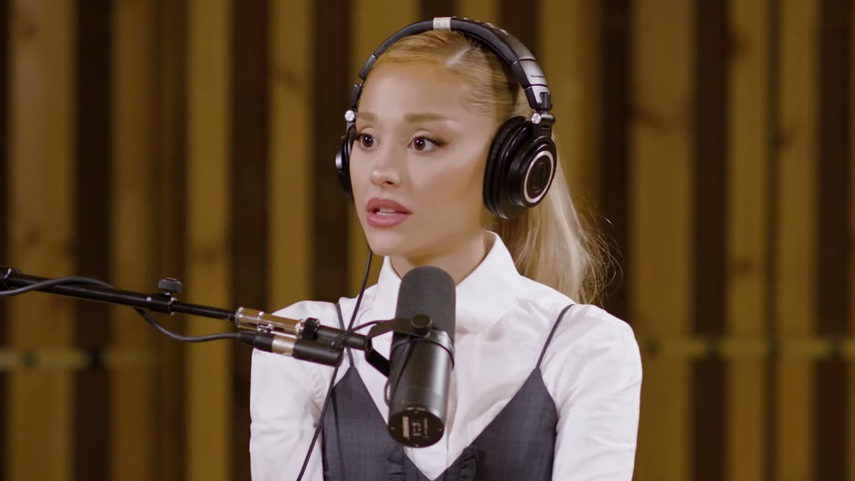 Ariana Grande wearing headphones and a white blouse layered with a dark vest, speaking into a microphone in a recording studio