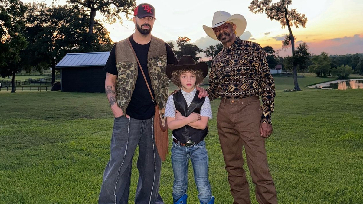 Drake, his son Adonis, and Dennis Graham are standing together outdoors. Drake and Dennis wear cowboy hats, while Adonis is in a cowboy outfit with blue boots