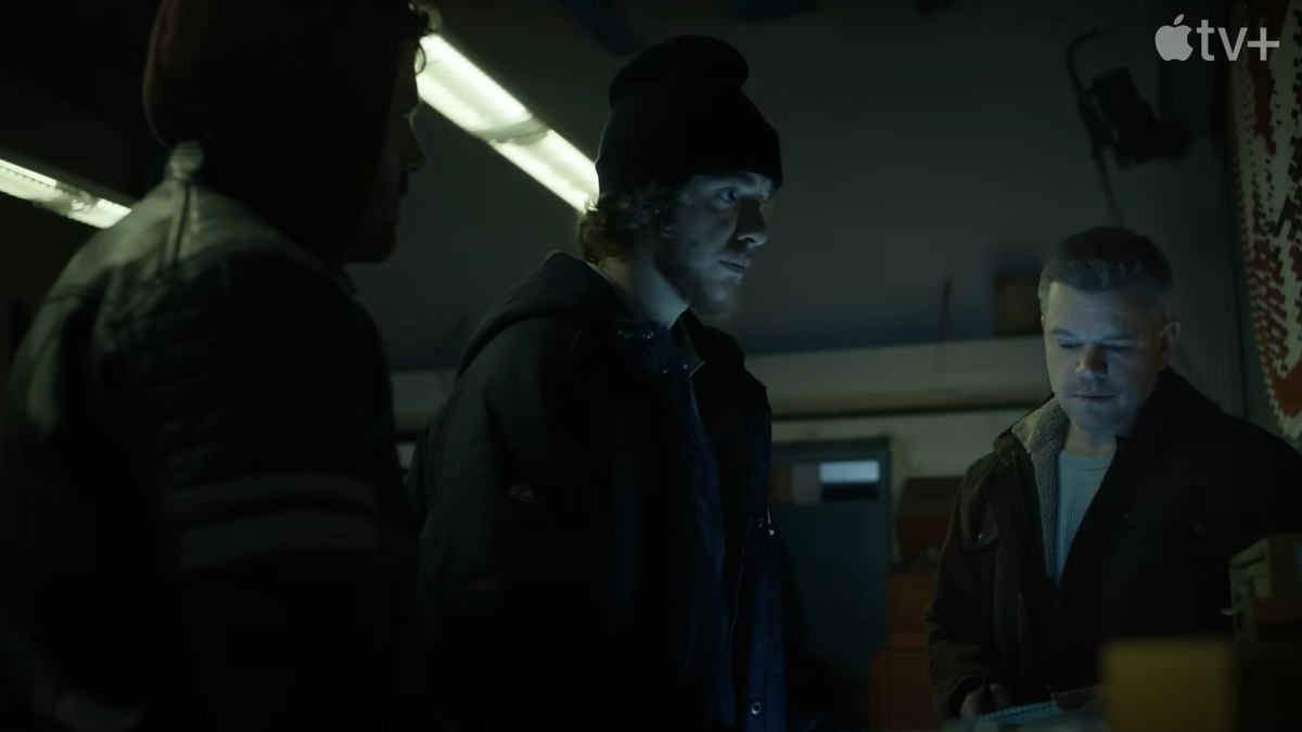 Three people, one in a beanie hat, another in a hood, and a third reading a document, are engaged in a tense conversation in a dimly lit room. Apple TV+ logo in corner