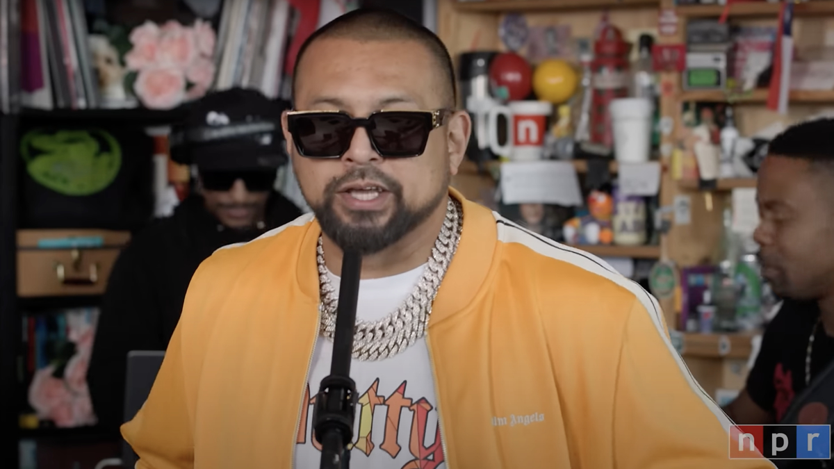 Sean Paul performs at NPR Tiny Desk Concert, wearing sunglasses and a yellow jacket with gold chains. Two musicians accompany him in the background