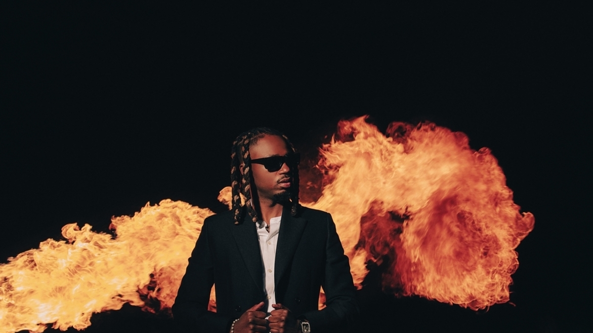 Musician in a dark suit and sunglasses stands confidently in front of large flames, creating a dramatic backdrop