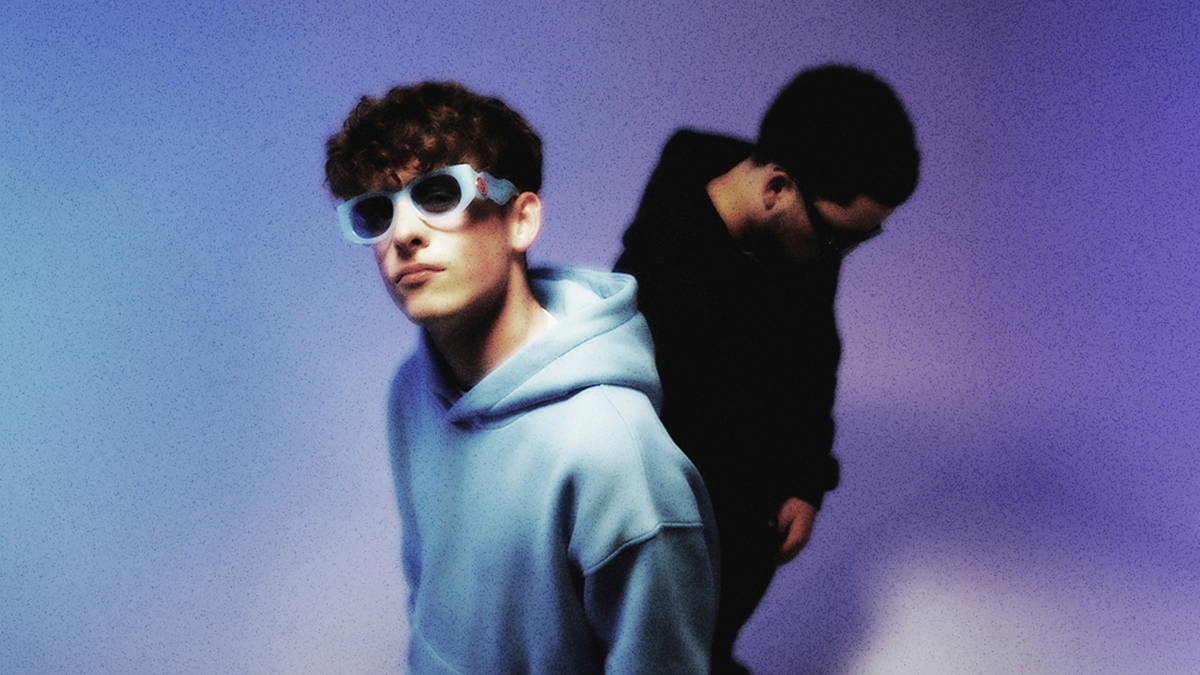 Two people in a music-themed image. One, in focus, wears a hoodie and large sunglasses; the other, blurred, stands in darker clothing