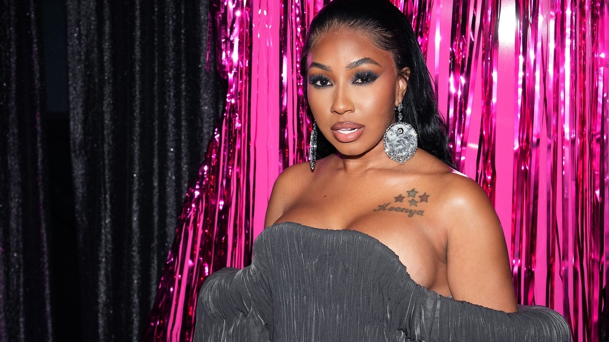 Yung Miami on a pink tinsel backdrop wearing an off-shoulder, pleated dress with large silver hoop earrings. She has a tattoo of stars and 'Zykojor' on her chest