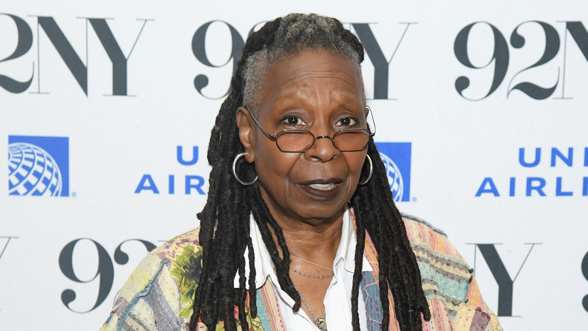 Whoopi Goldberg in a patterned jacket and glasses, standing in front of a logo wall