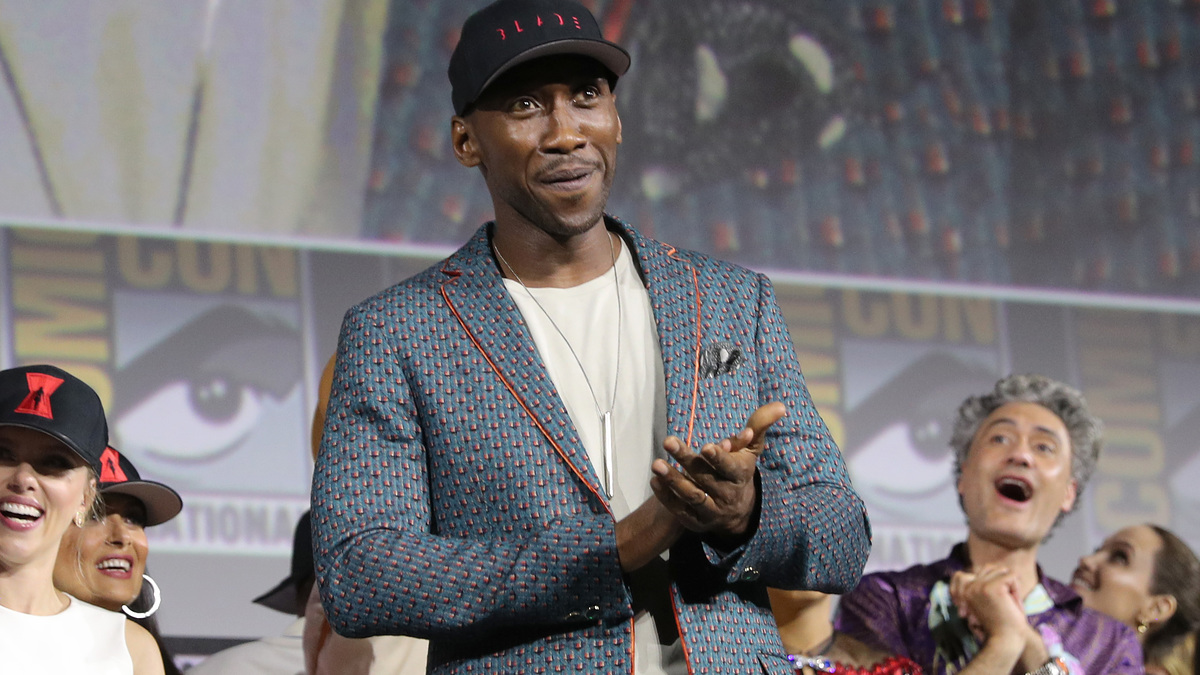 Mahershala Ali on stage at Comic-Con, wearing a patterned blazer and a black hat. Other attendees are visible in the background