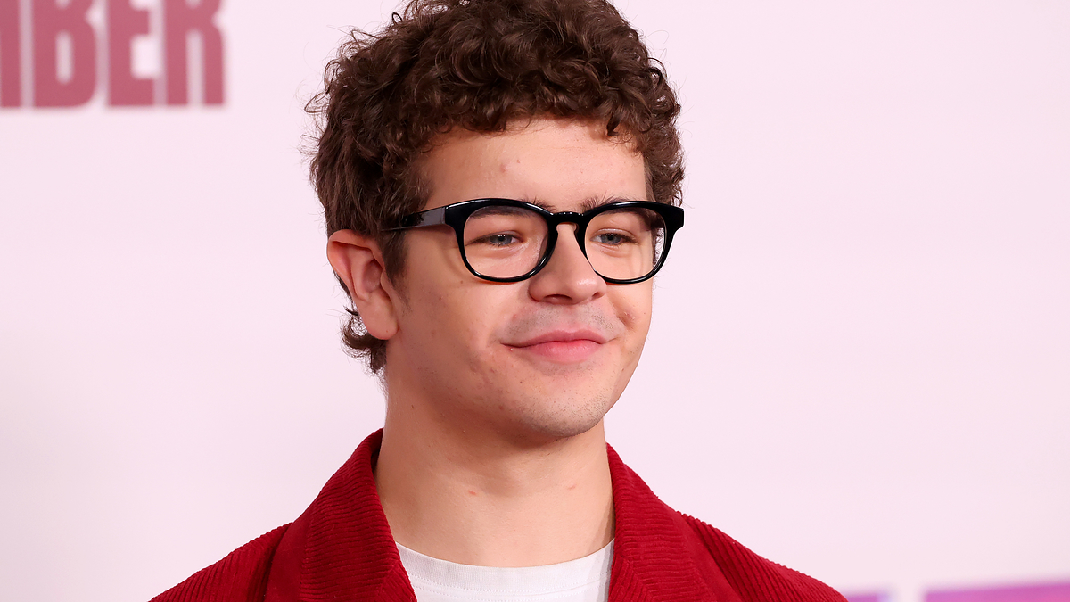 Gaten Matarazzo at an event, wearing black-frame glasses, a white shirt, and a casual red jacket