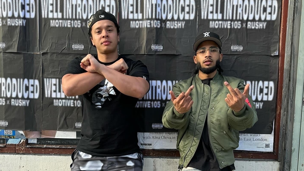 Motive105 and Rushy stand in front of posters for their music collaboration "Well Introduced." Motive105 crosses his arms, while Rushy shows two middle fingers