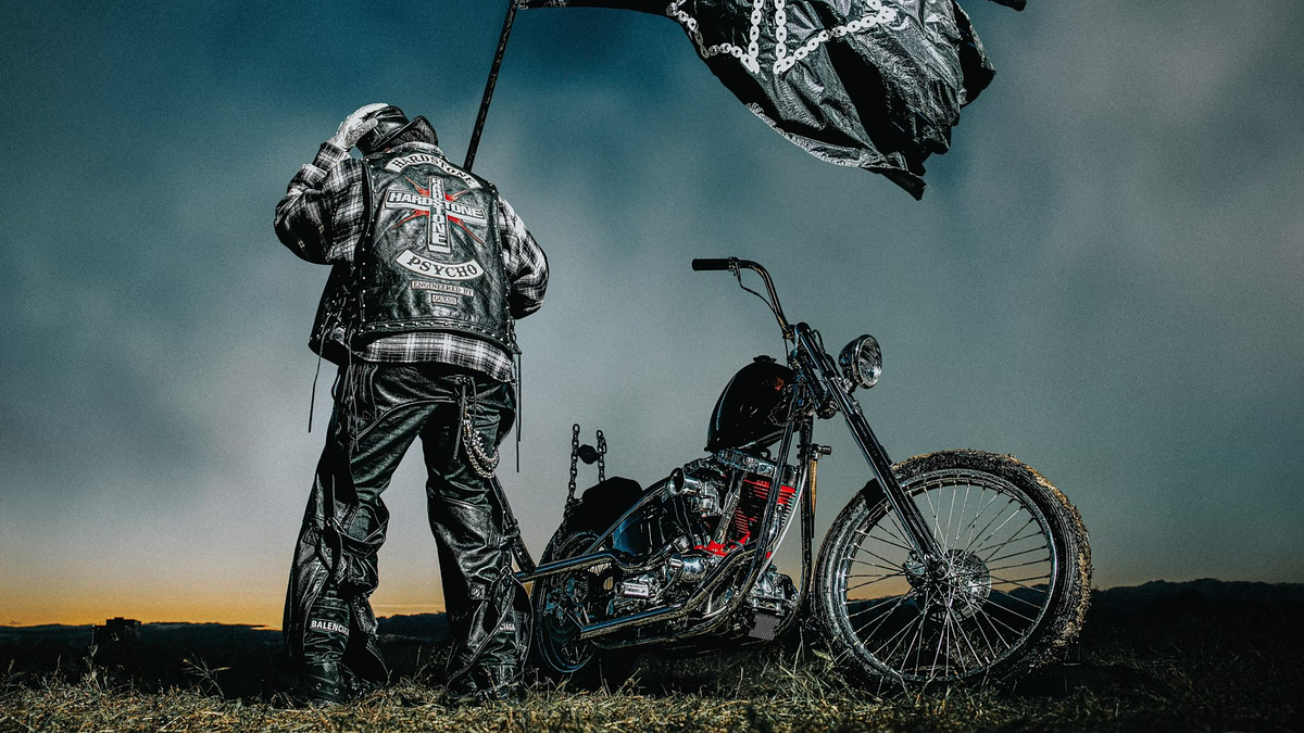 A biker in leather gear stands by a custom motorcycle, holding a flag. The back of the biker's jacket reads "Hardcore Carnivore Psycho." The image is related to music