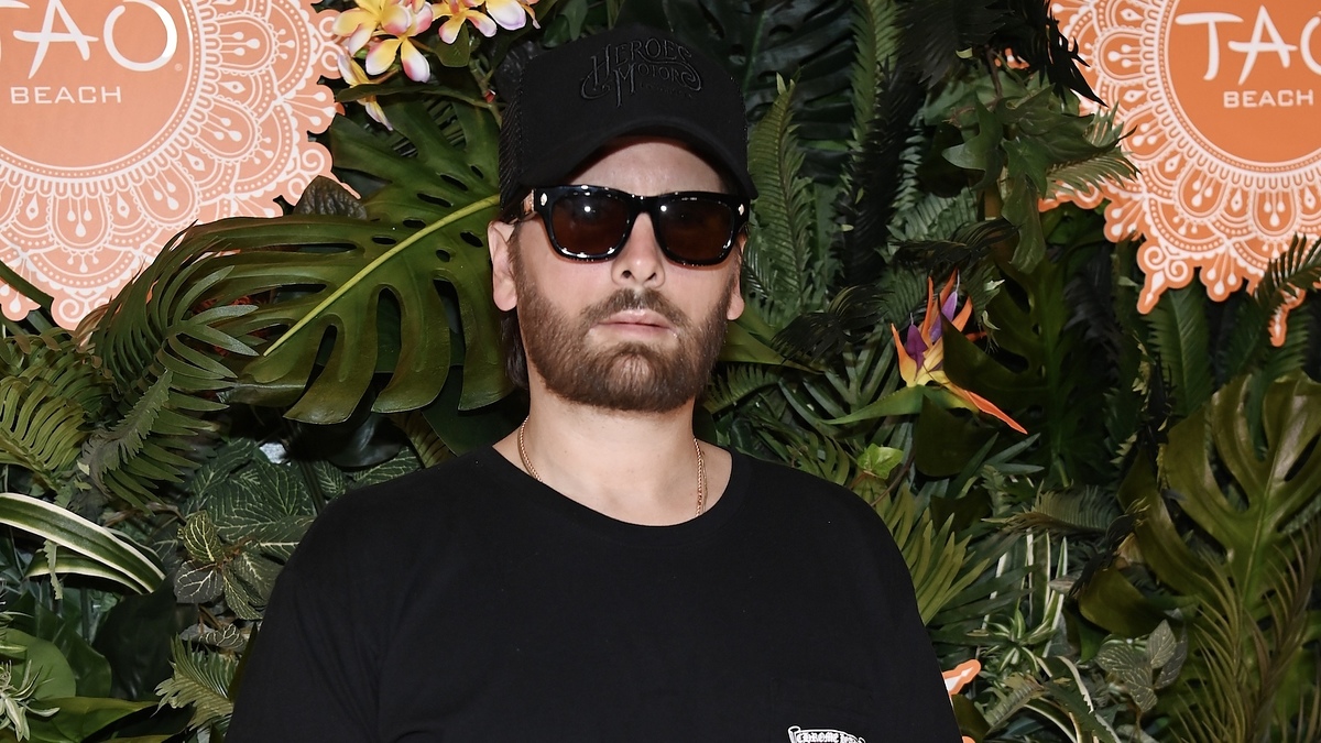 Scott Disick wearing a black t-shirt and sunglasses, poses in front of a tropical-themed backdrop at the Tao Beach event