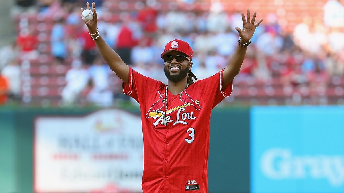 A person wearing a St. Louis Cardinals baseball uniform and cap smiles and raises both arms while holding a baseball