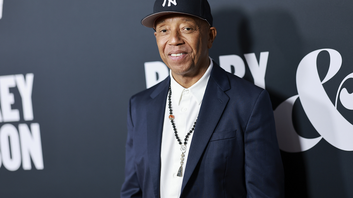 Russell Simmons at an event, wearing a navy suit, white shirt, black necklace, and a black New York Yankees cap