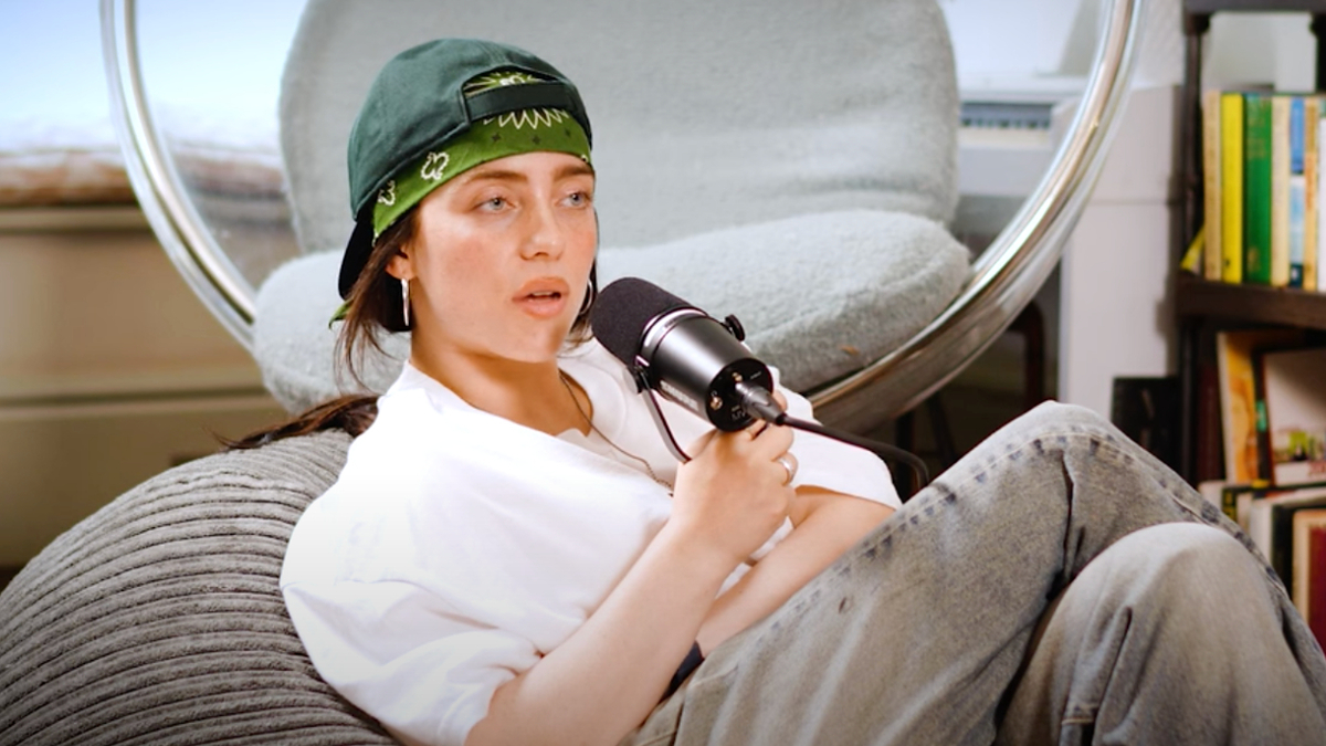 Billie Eilish is lounging in a chair, wearing a casual white shirt and gray pants, with a green bandana and hat, holding a microphone in a recorded interview