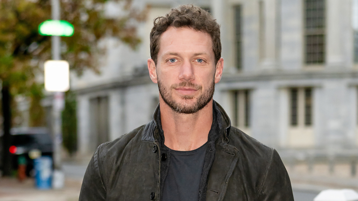 A person with short, curly hair and a beard, wearing a black jacket and dark shirt, standing on a city street with buildings in the background. Names unknown