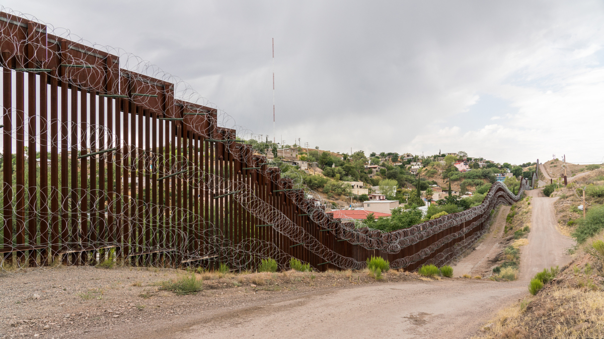 A border fence topped with barbed wire extends across hilly terrain, with a dirt road and scattered houses in the background under a cloudy sky