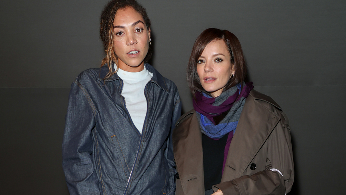 Two women, one in a denim suit holding a plaid purse, and the other in a trench coat with a fur bag, pose together against a plain backdrop. Names unknown