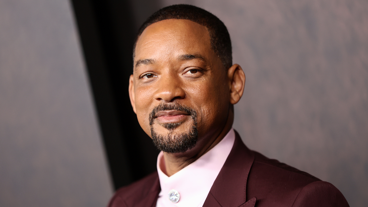 Will Smith is smiling, wearing a formal suit with a white shirt and a decorative button