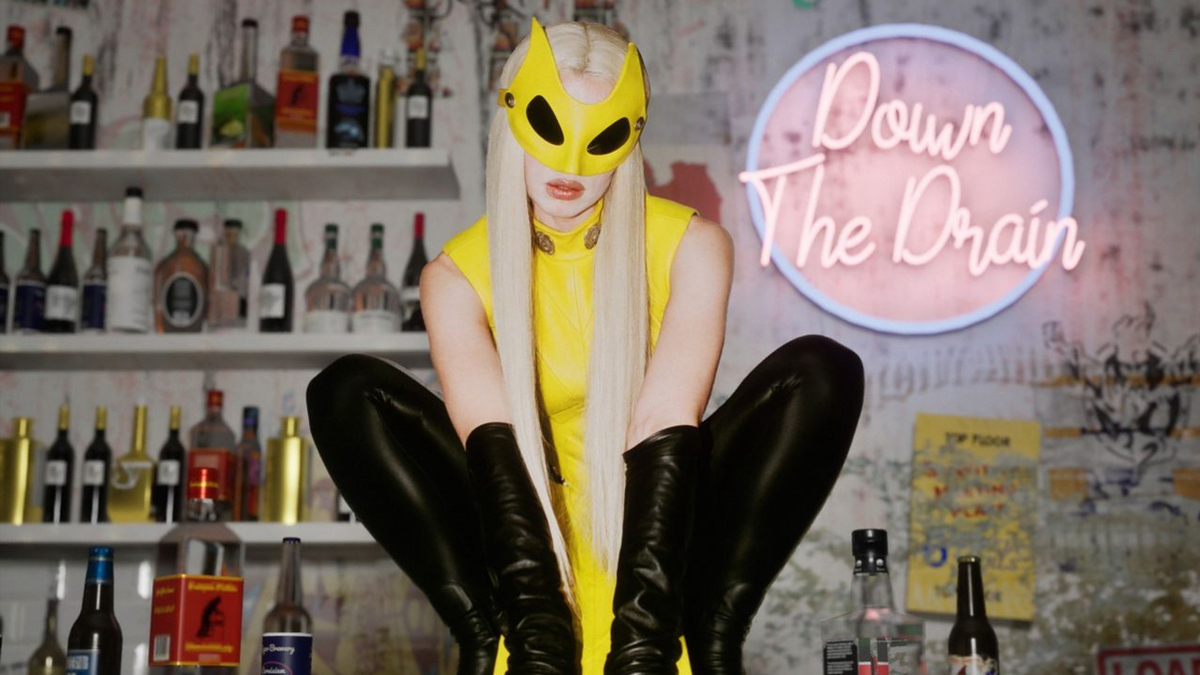 Person in yellow mask and long gloves sitting with neon sign "Down The Drain" and bottles in background