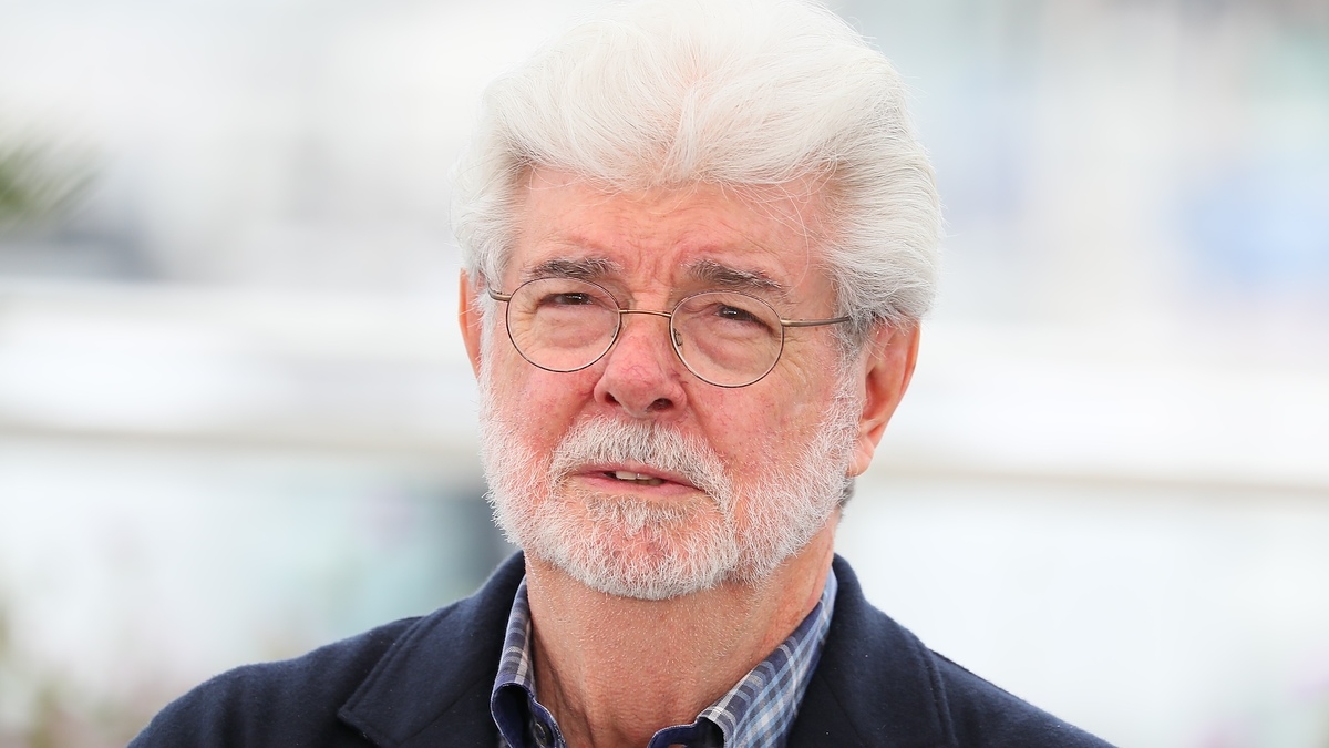 An older man with glasses and a beard is wearing a dark jacket and a checkered shirt, looking into the distance