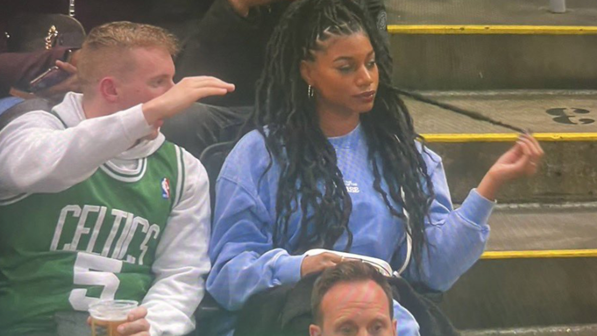 A man wearing a Celtics jersey and holding a drink sits next to a woman in a blue sweater with long braided hair as they watch a sports event