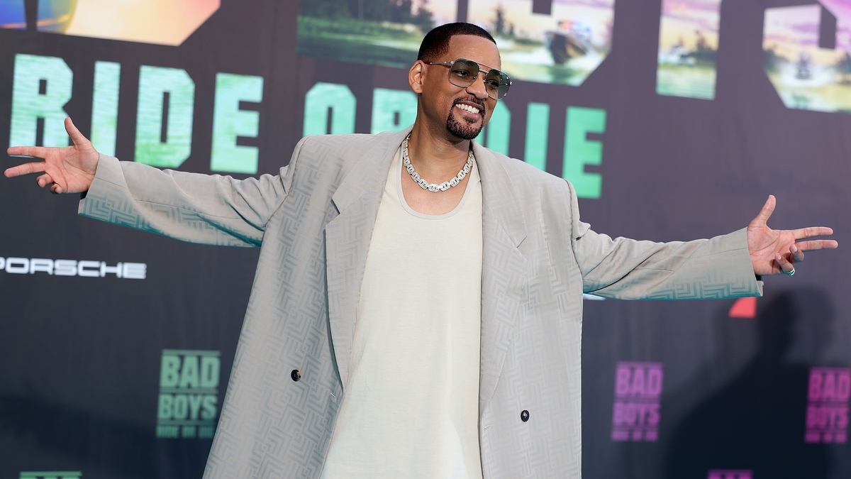 Will Smith at a movie premiere wearing a light-colored oversized suit, a white T-shirt, sunglasses, and a chain necklace, standing with arms spread open